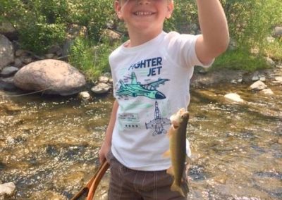 Young smiling Boy standing in a river holding up a cutthrroat trout in one hand and a fishing net in the other - Lazy L&B Dude Ranch Wyoming